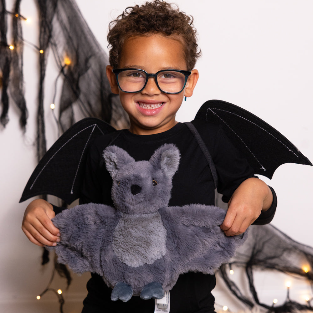 Child holding a plush gray bat toy with outstretched wings, smiling and wearing glasses.