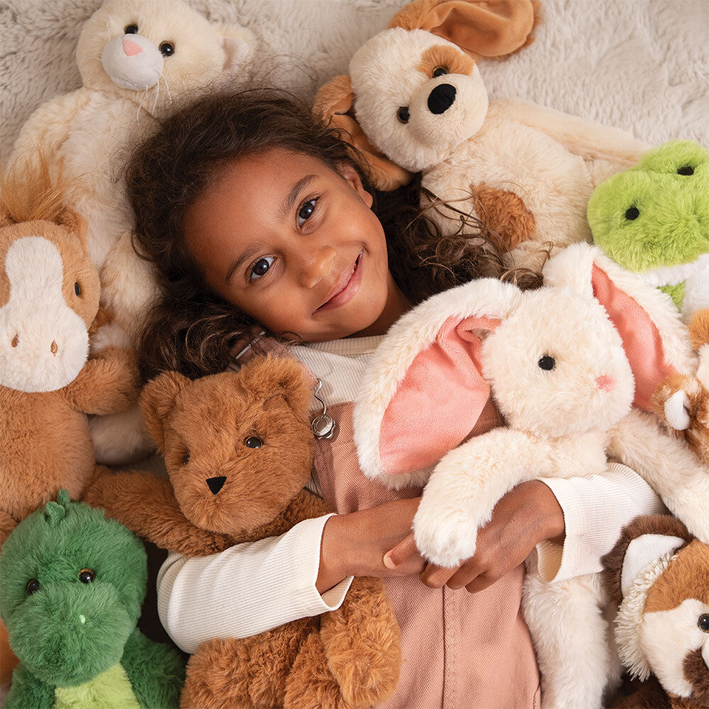 Smiling child cuddling soft plush puppy and various animal stuffed toys on white fuzzy blanket.