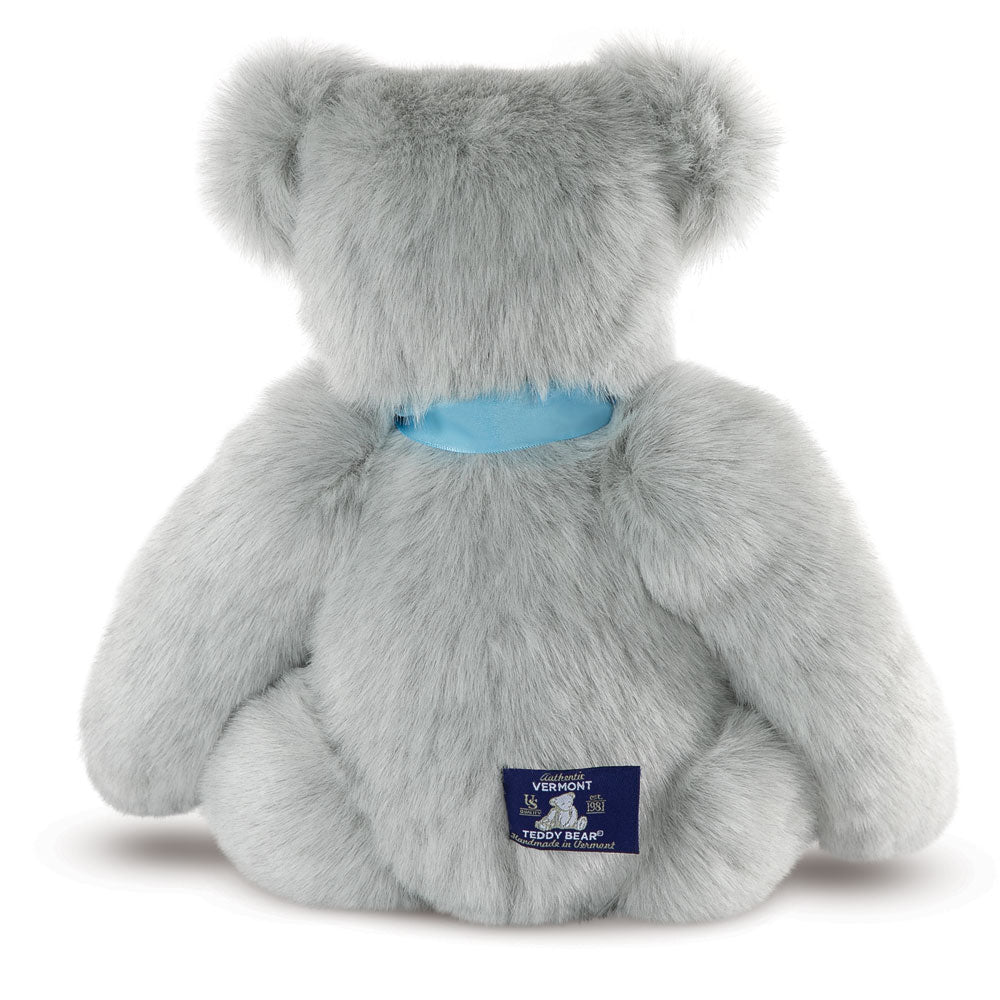 Grey plush teddy bear with blue satin ribbon around neck, sitting facing backward.