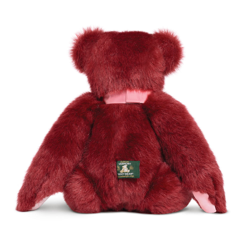 Plush deep red teddy bear with a pink satin neck ribbon and a green label on the back.