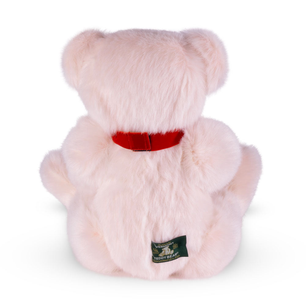 Soft pink plush teddy bear with red ribbon collar and green fabric tag on back.