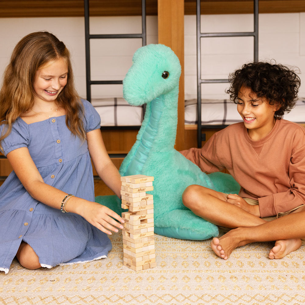 Large plush turquoise dinosaur with long neck, sitting between two children playing Jenga on a beige patterned rug.