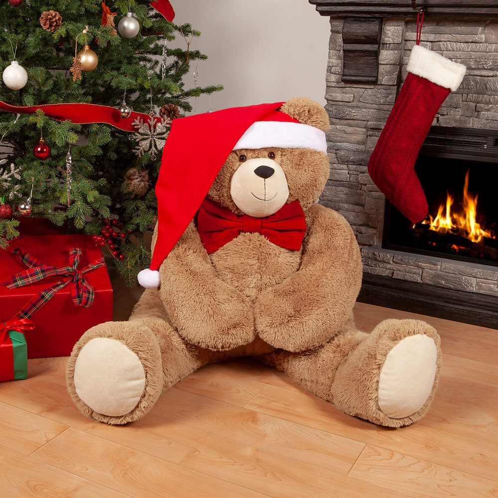 4 ft brown plush bear wearing red bow tie and long Santa hat sitting by decorated Christmas tree and fireplace.