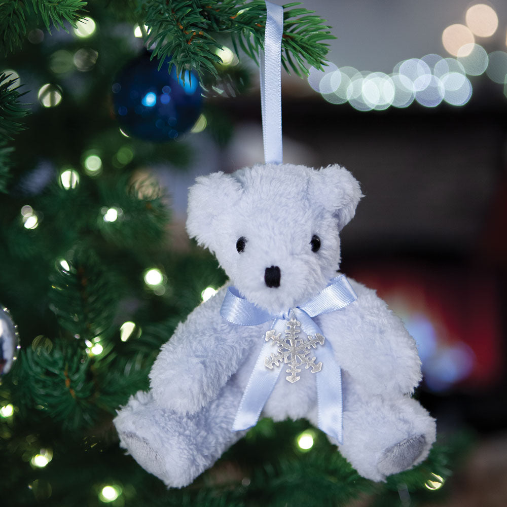 Plush white teddy bear ornament with blue satin ribbon and silver snowflake charm hanging on Christmas tree.