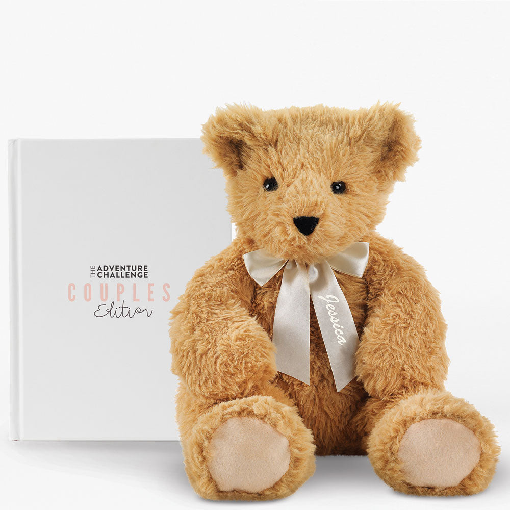 Soft light brown teddy bear with beige paws and a satin ribbon bow, sitting in front of a white box labeled Adventure.