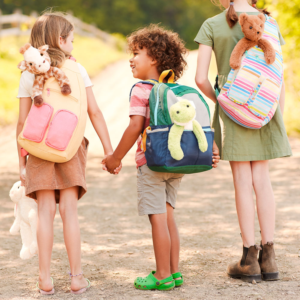 Three children wearing colorful backpacks with plush animal pals attached on a sunny outdoor path.