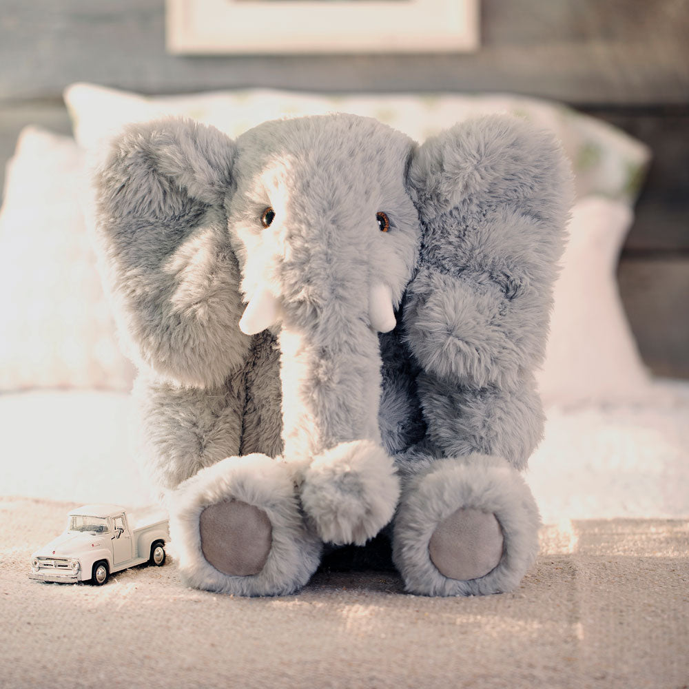 Gray plush elephant with large floppy ears and soft texture sitting on a bed next to a small white toy car.