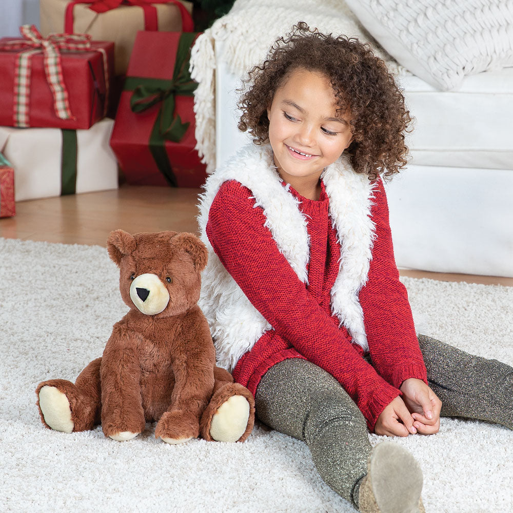 Brown plush teddy bear with cream snout and paw pads sitting on carpet next to girl in red long-sleeve and white fuzzy vest.