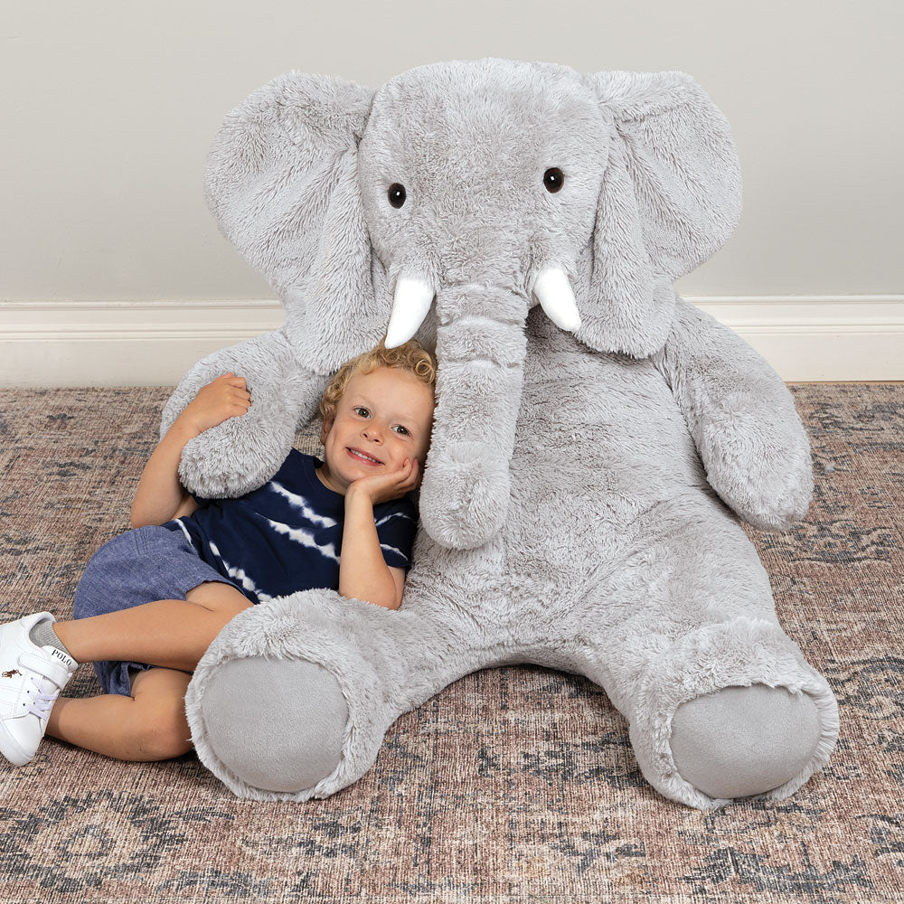 Giant 4 ft. plush gray elephant with white tusks and big floppy ears hugging young child on rug.