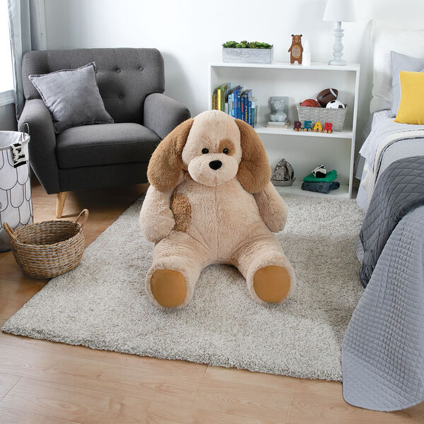 Plush tan and brown stuffed puppy sitting on a beige rug in a cozy bedroom setting.