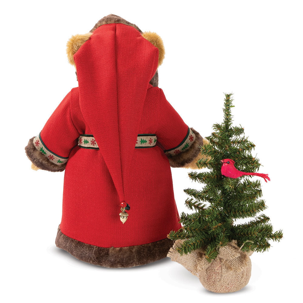 Plush bear dressed in red Santa cloak with fur trim, standing next to small pine tree with red bird ornament.
