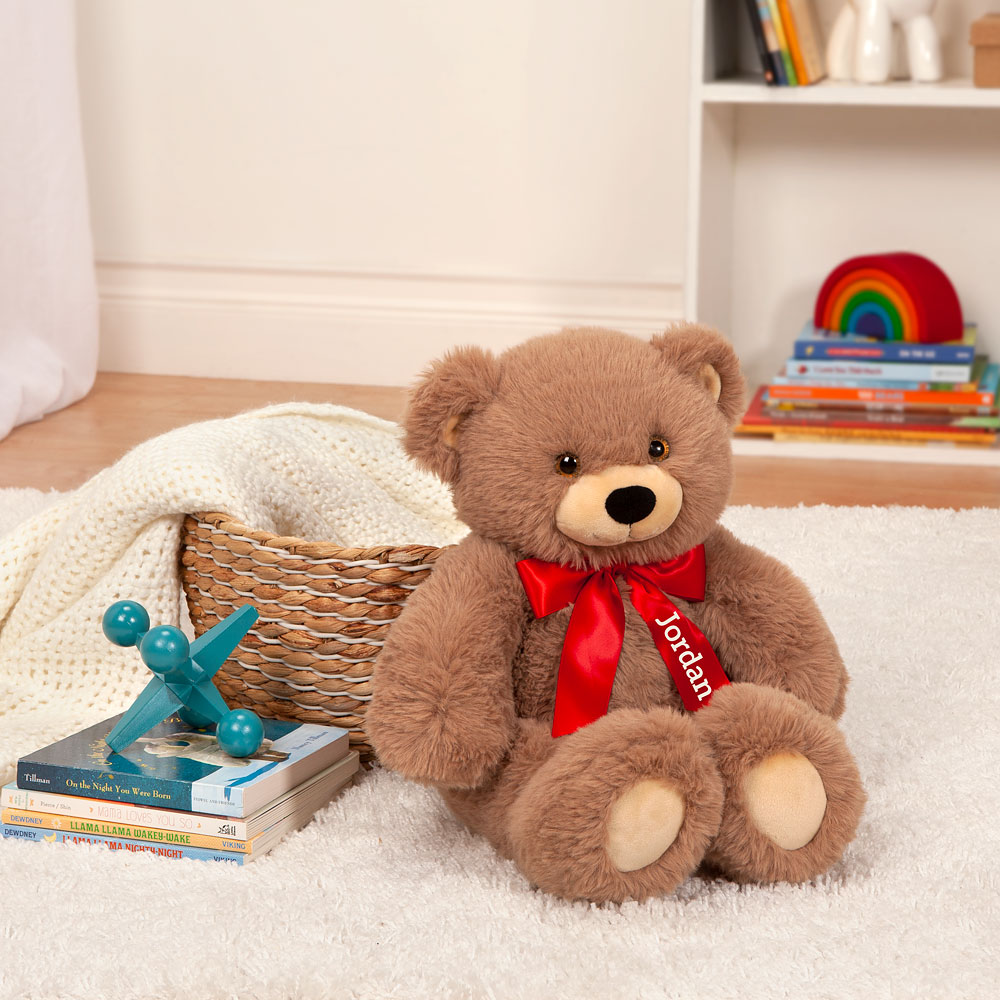 Brown teddy bear with red satin bow sitting on white rug near wicker basket and books.
