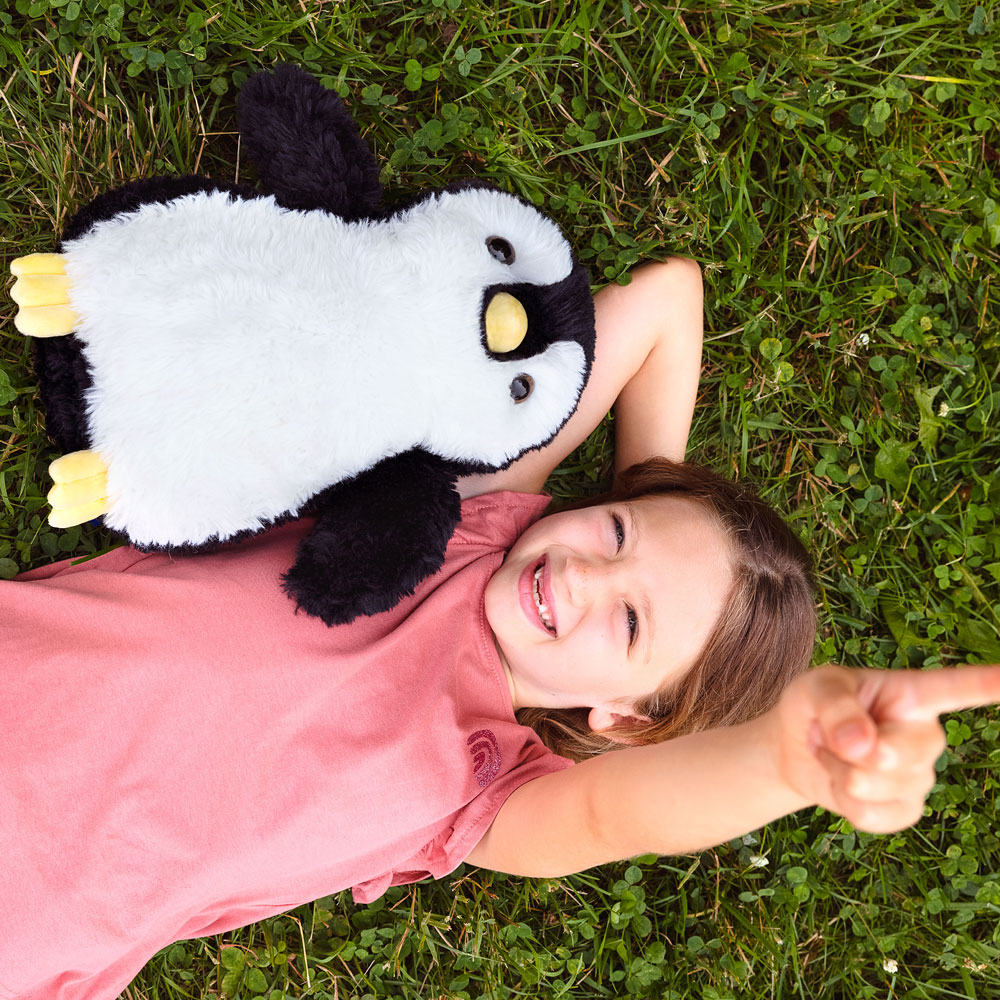 White and black plush penguin with yellow feet and beak held by smiling girl in pink shirt on grass.