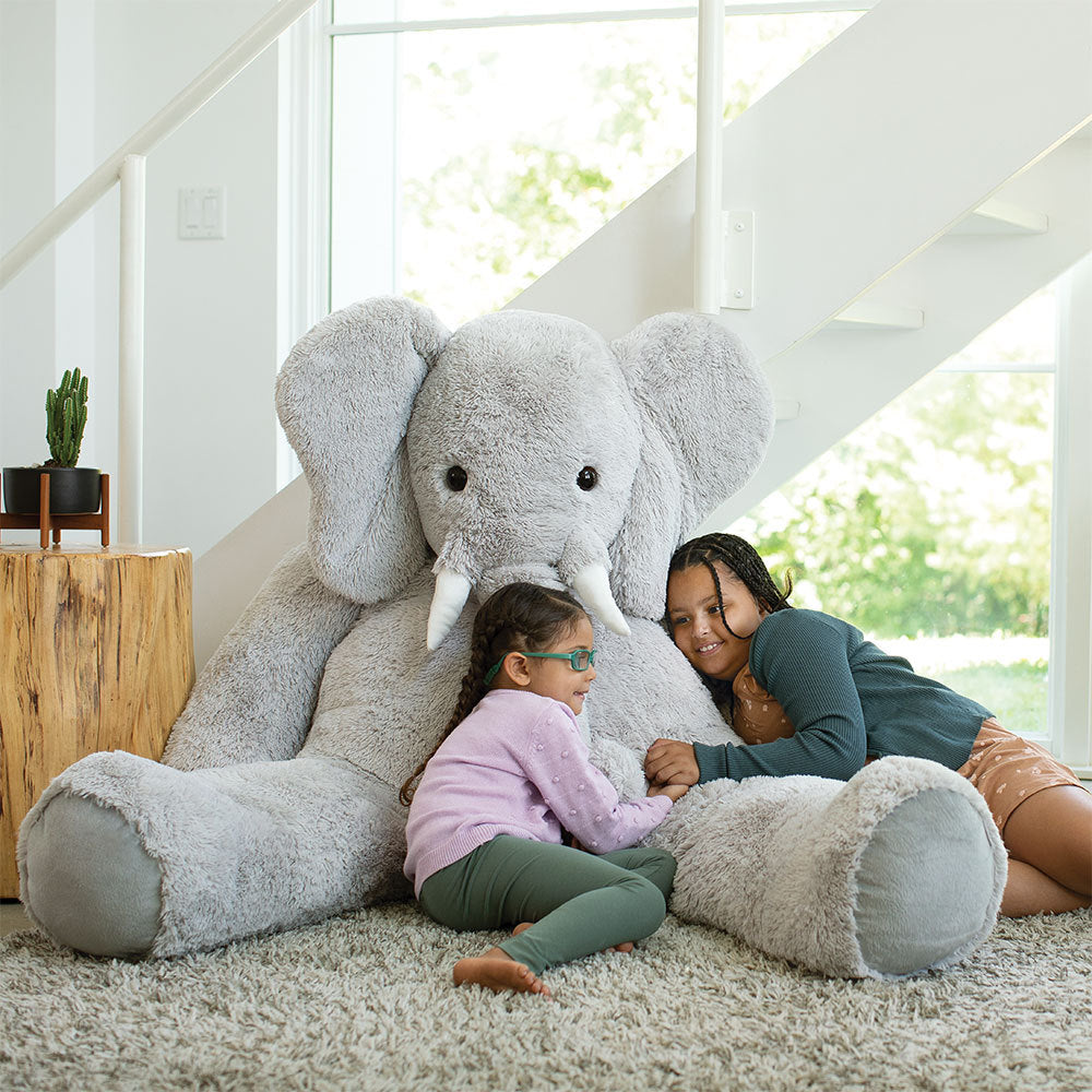 6 ft. gray plush giant elephant with large ears and trunk, two kids hugging it indoors by stairs.