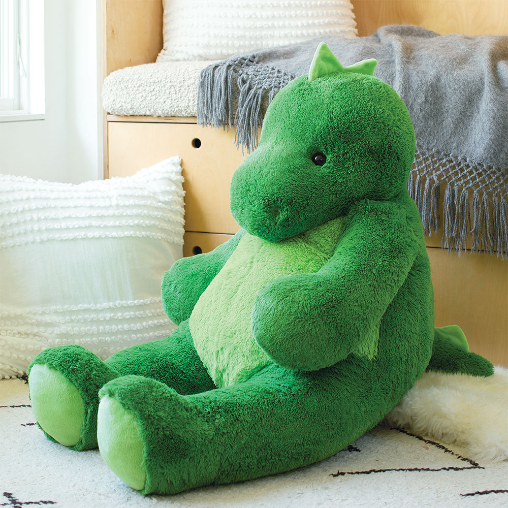 Large green plush dinosaur with light green belly and soft texture sitting on white rug near cushions and blanket.