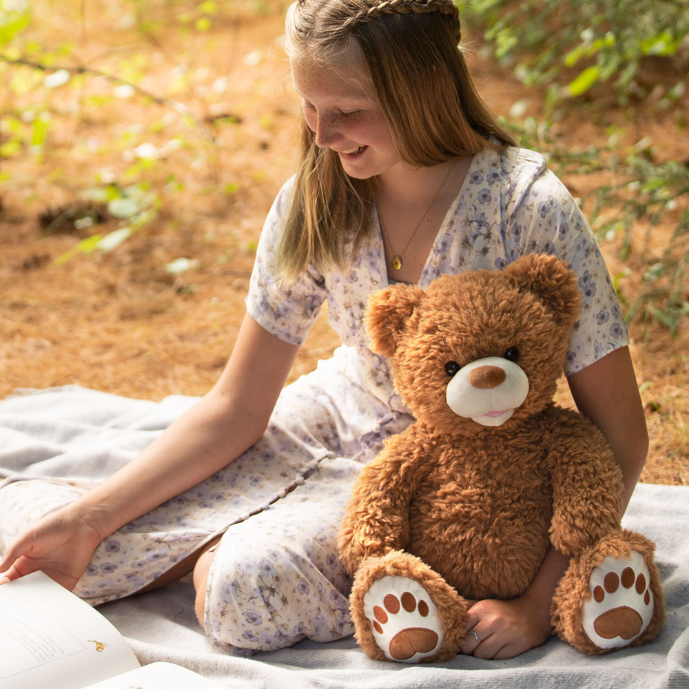 Brown fuzzy teddy bear with large paws and a white snout sitting on a blanket with a girl in a floral dress.