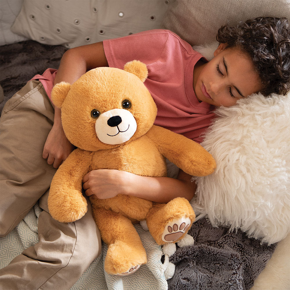 Child cuddling a 20-inch light brown teddy bear with embroidered face and paw details on a bed.