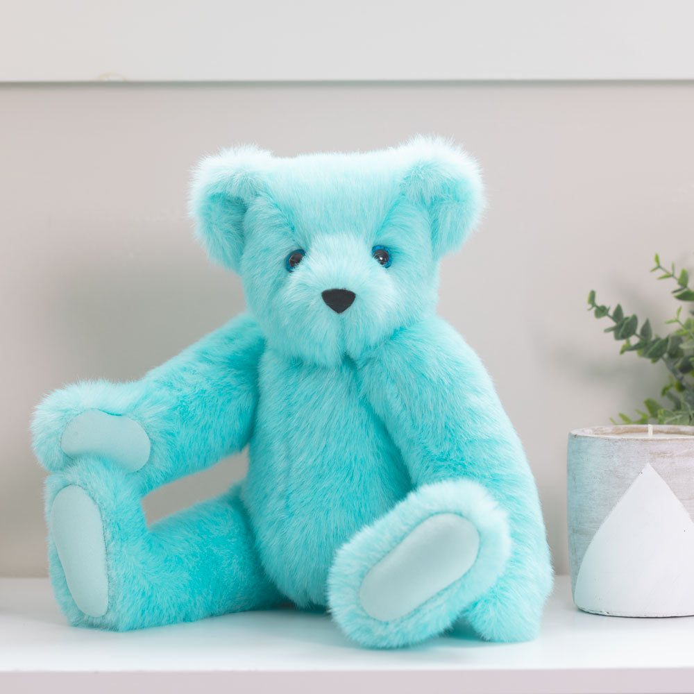 Soft blue teddy bear plush with white paw pads sitting next to a small white planter with greenery.