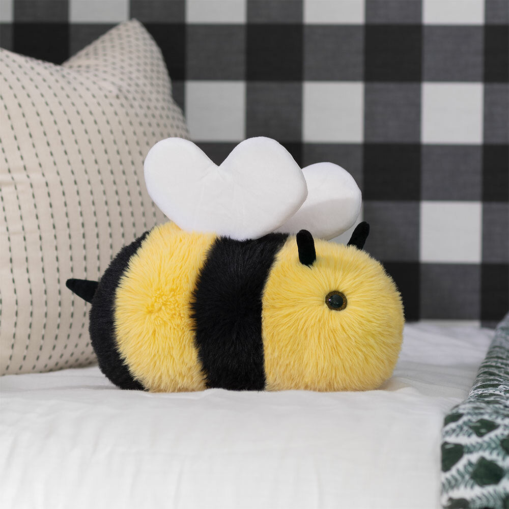 Soft yellow and black striped plush bee with white wings on bed with checkered pillow background.