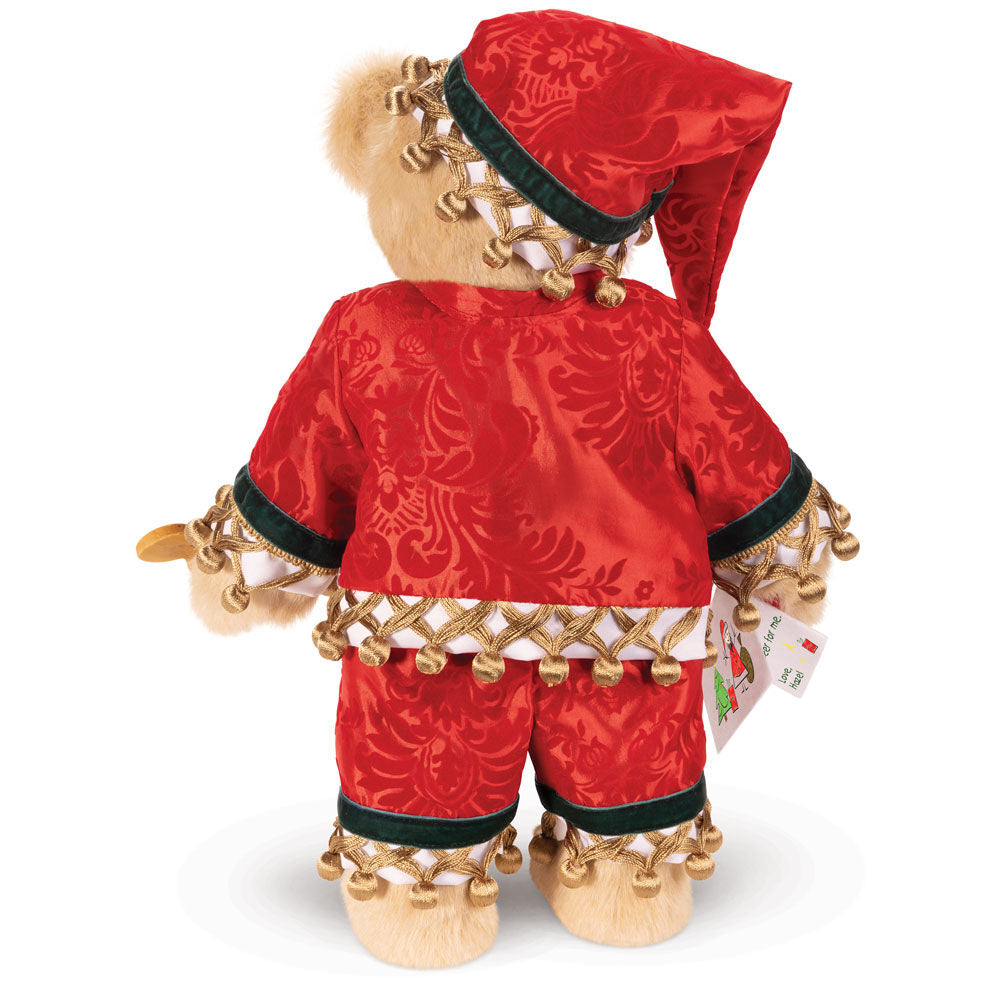 Plush bear dressed in red velvet Santa outfit with gold jingle bells and green trim, holding gift tag.