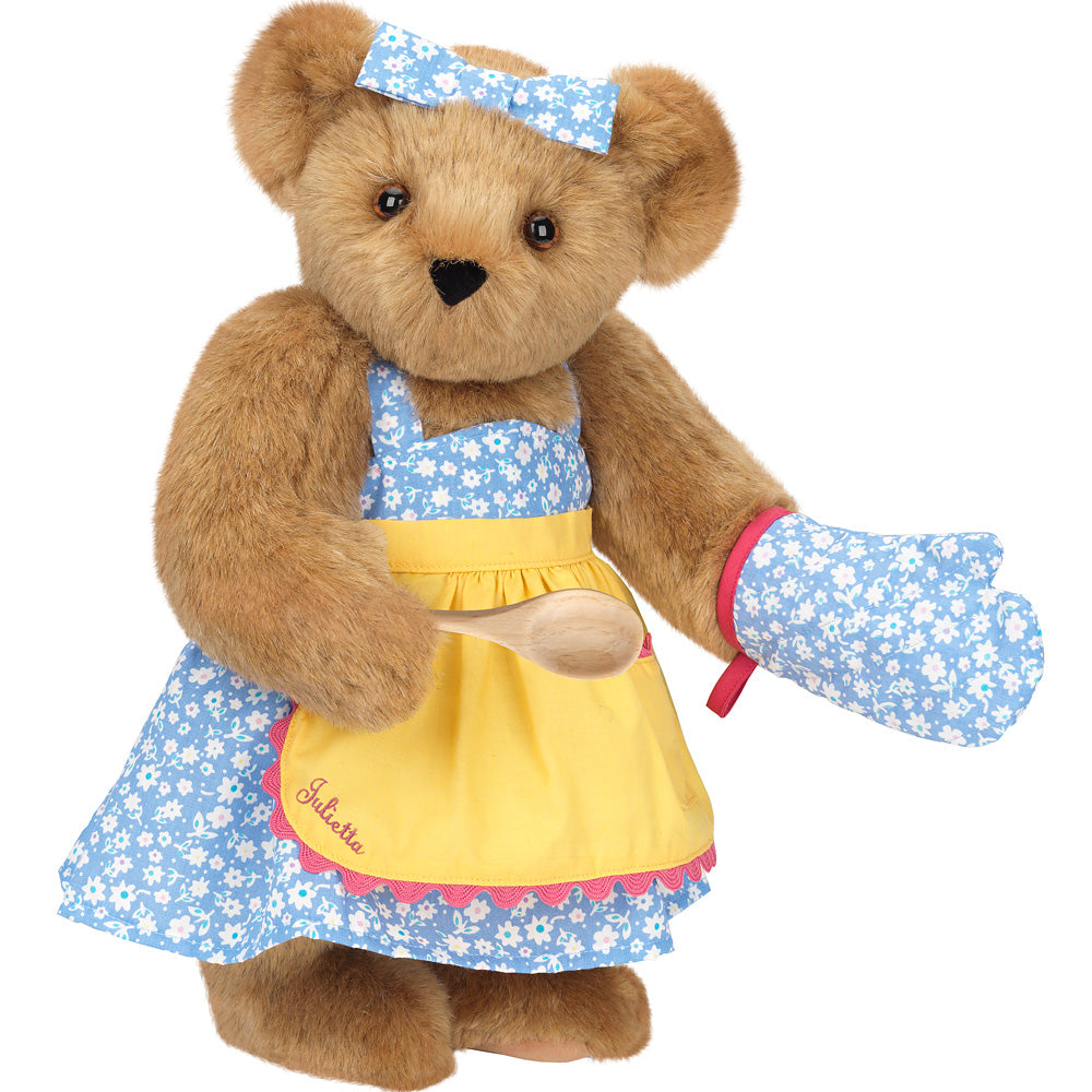 Brown teddy bear wearing blue floral apron, matching oven mitt, and yellow apron with wooden spoon.