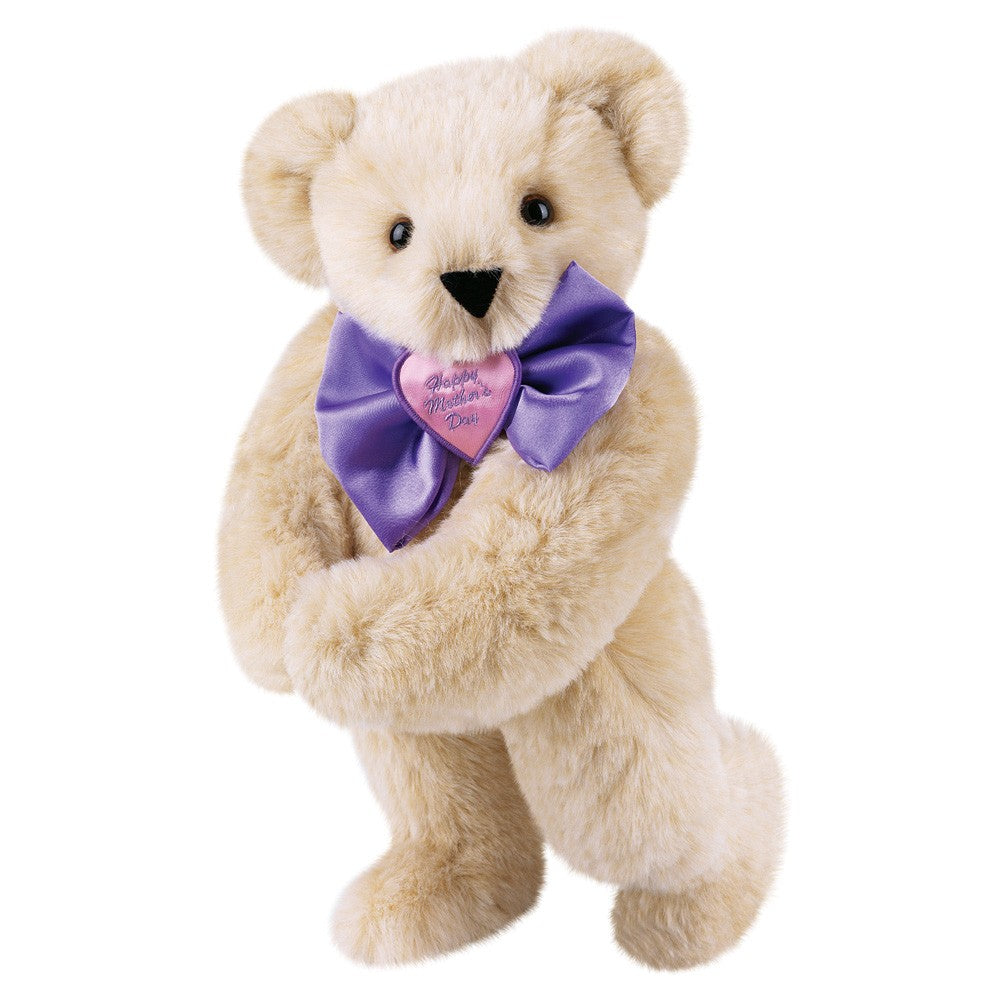 Cream-colored plush teddy bear standing with purple satin bow tie and heart-shaped pendant reading "Happy Mother's Day".