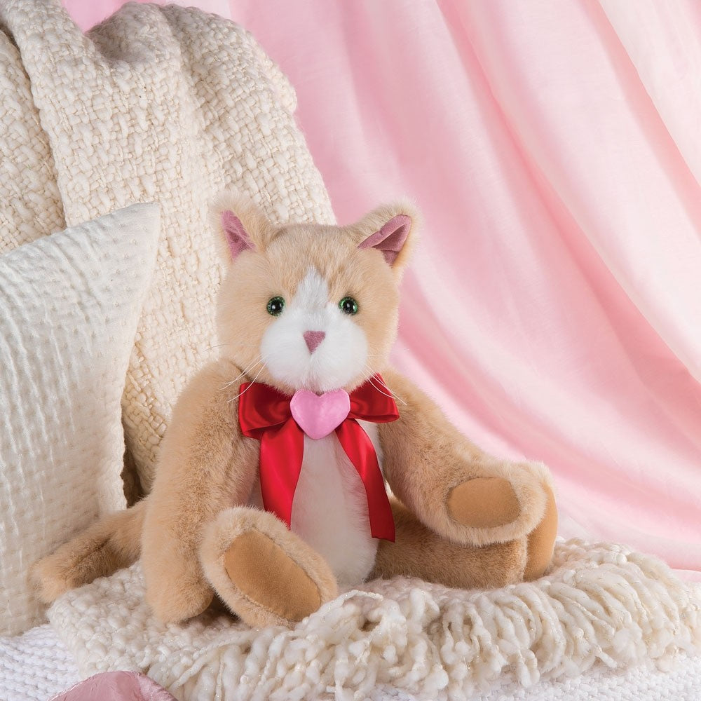 Plush tan and white cat with pink nose, red ribbon, and heart pendant sitting on beige knit blanket with pink backdrop.