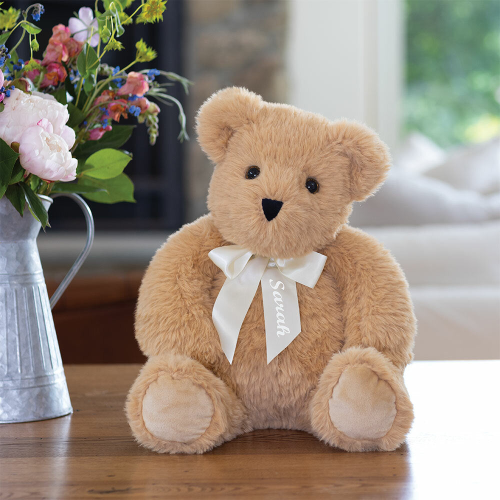 Plush light brown teddy bear with a cream bow sitting on a wooden surface near a floral arrangement.