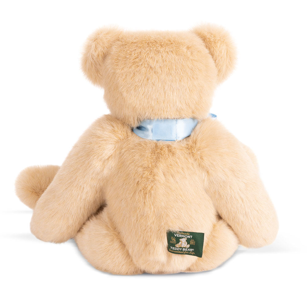 Light tan plush teddy bear with seated pose, wearing a shiny blue ribbon around its neck and a green tag on its back leg.