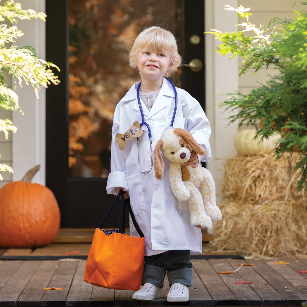 A young child dressed as a doctor stands on a wooden porch holding an orange tote bag and a plush dog toy. The child wears a white lab coat, stethoscope, and dark pants, with pumpkins, hay bales, and greenery around them.