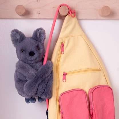 Gray plush bat toy hugging pink and yellow fabric pouch with front pockets and hanging loop.