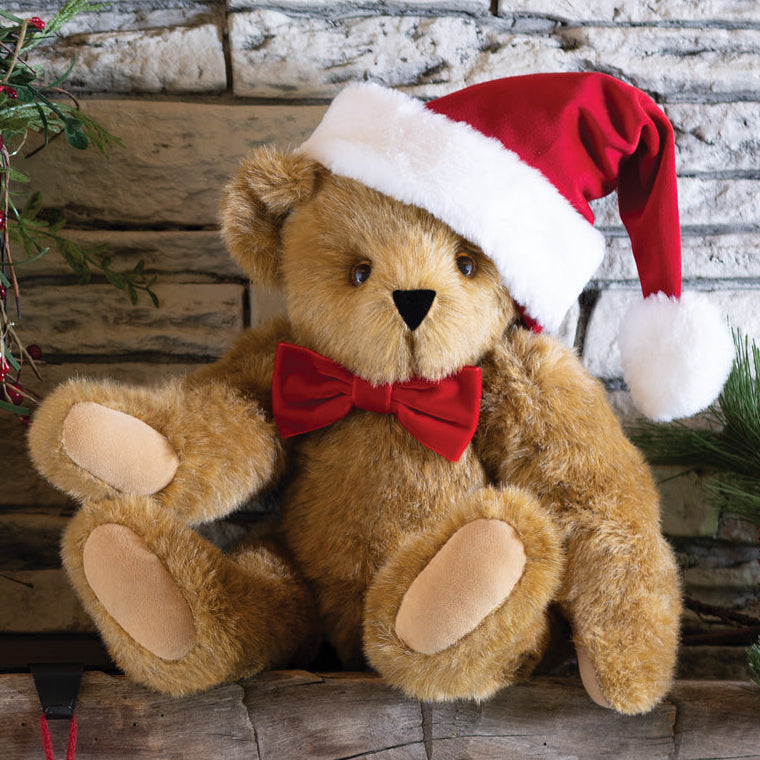 Brown plush teddy bear wearing a red Santa hat and red bow tie sitting on a stone ledge by holiday greenery and pinecones.