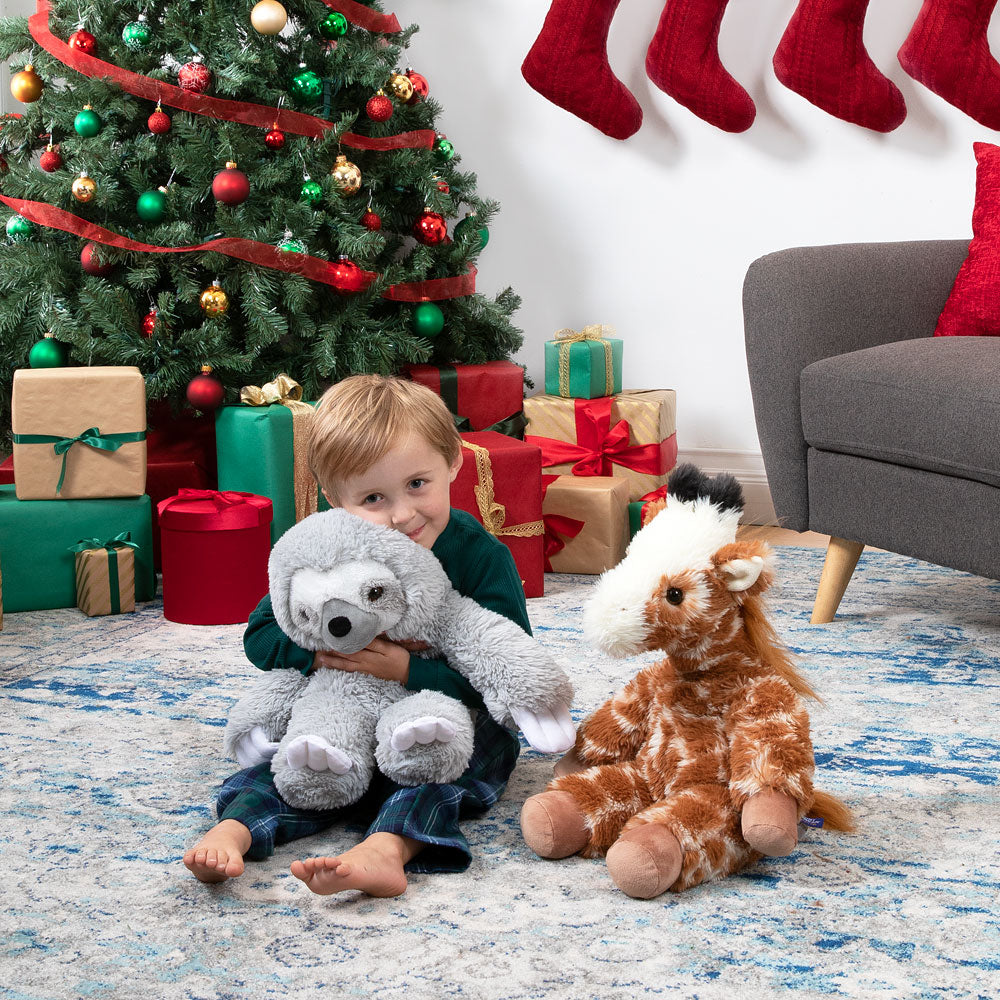 Child holding a stuffed sloth toy in a festive room with Christmas tree and presents and a Giraffe stuffed animal.
