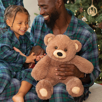 Man and child in plaid pajamas holding a teddy bear with a Christmas tree in the background