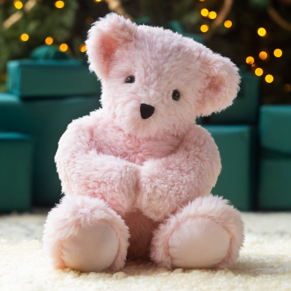 Pink teddy bear sitting on a light surface with blurred green and gold festive background