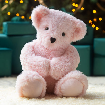 Pink teddy bear sitting on a light surface with blurred green and gold festive background