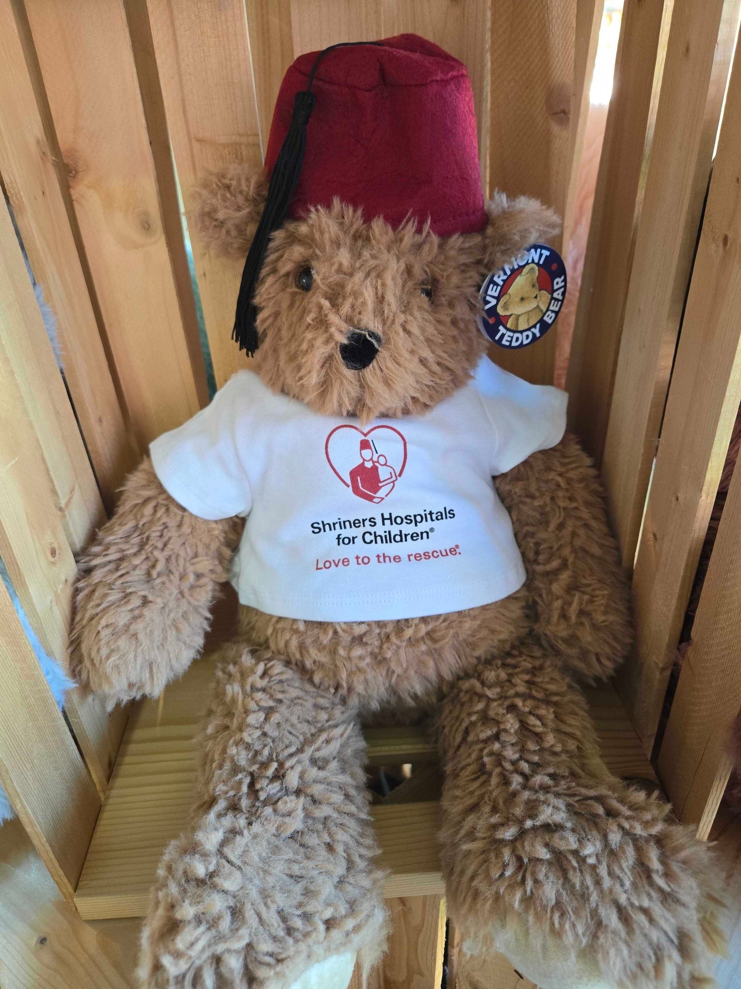 Brown teddy bear wearing a red fez and white shirt with St. Jude logo and text sitting on wooden shelf.