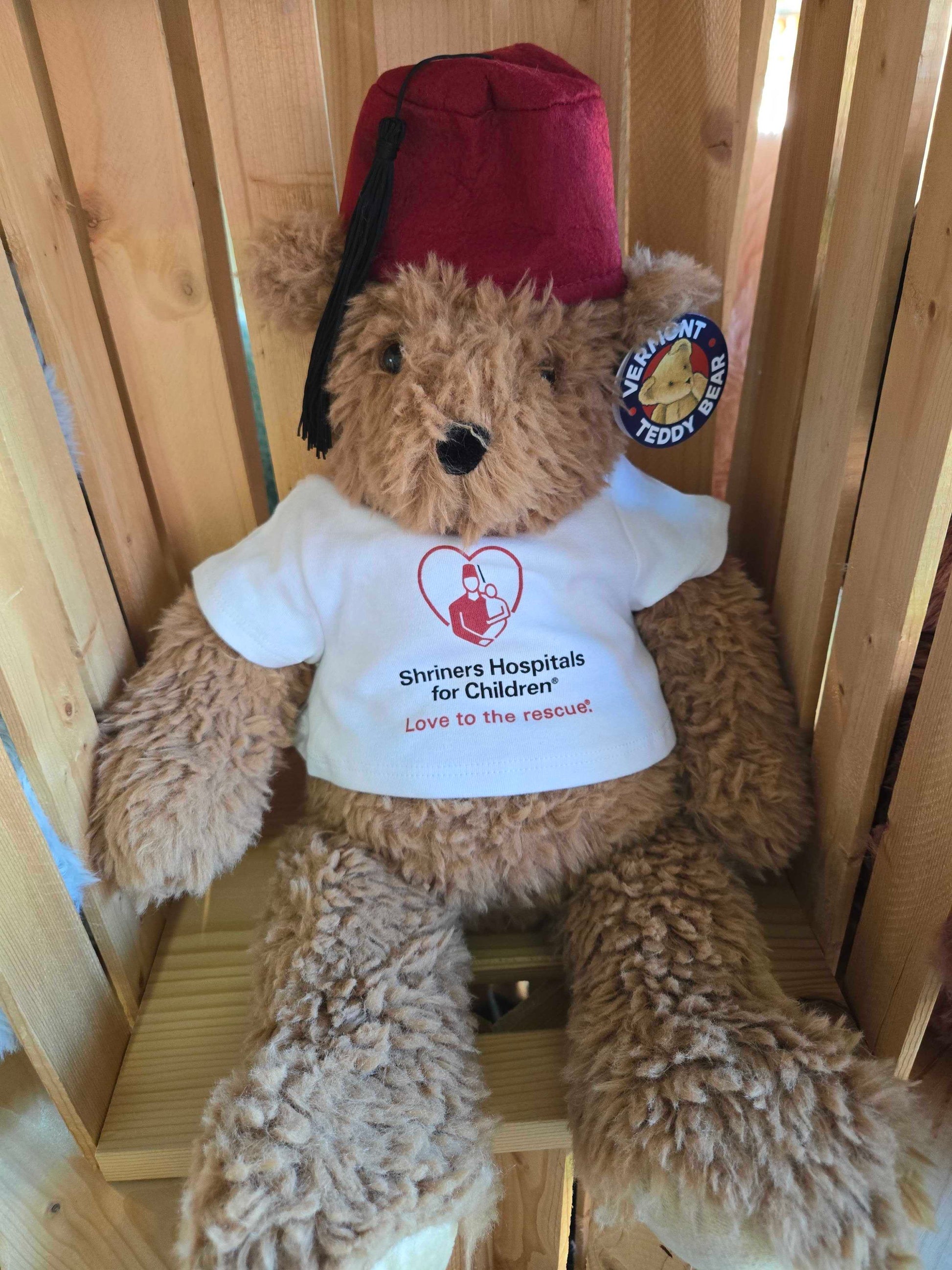 Brown teddy bear wearing a red fez and white shirt with St. Jude logo and text sitting on wooden shelf.