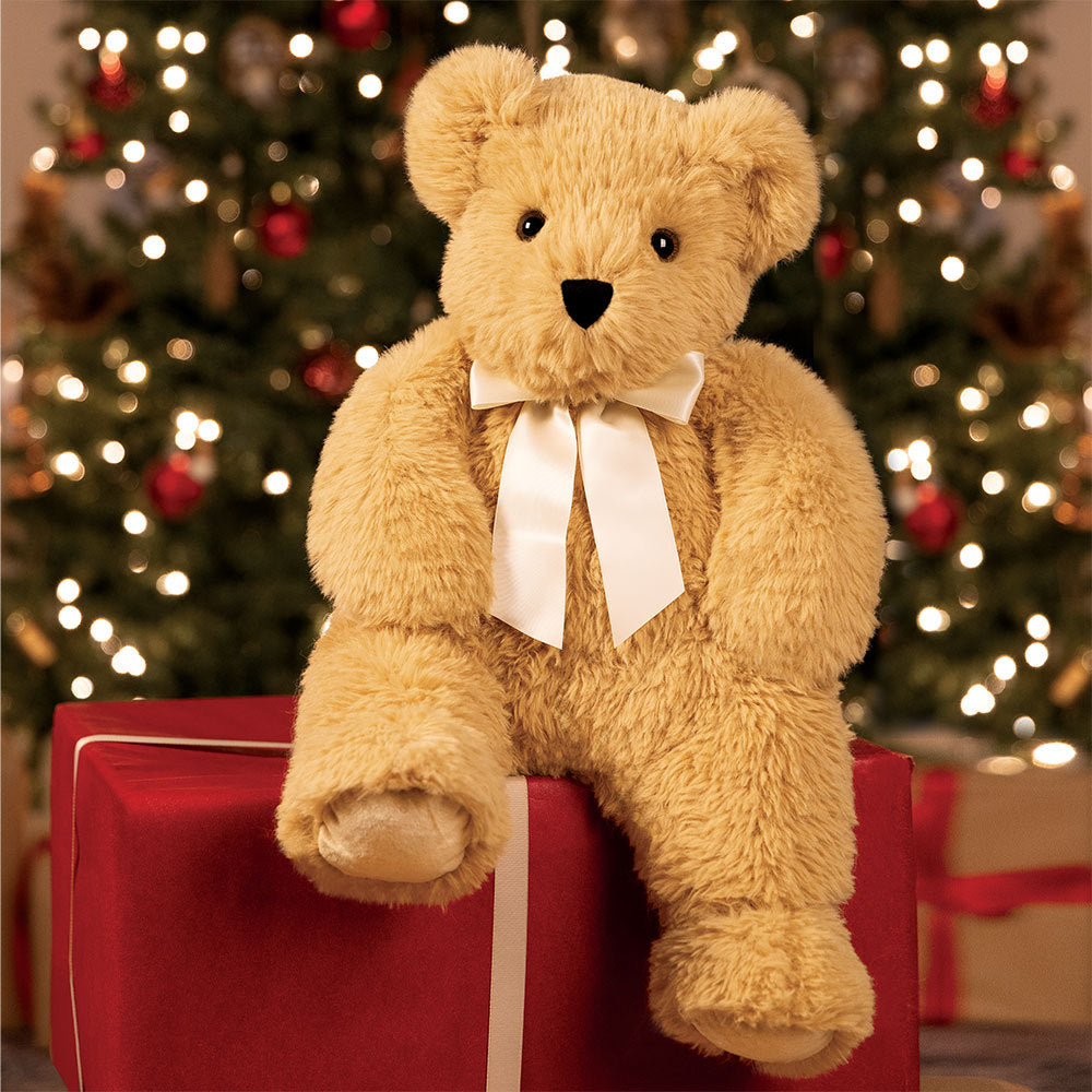 Plush teddy bear with a white bow sitting on a red gift box in front of a decorated Christmas tree.