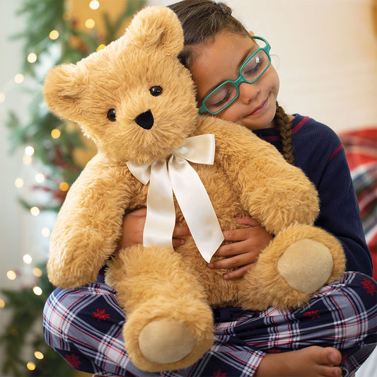 Child holding a teddy bear with a white ribbon in front of a Christmas tree.