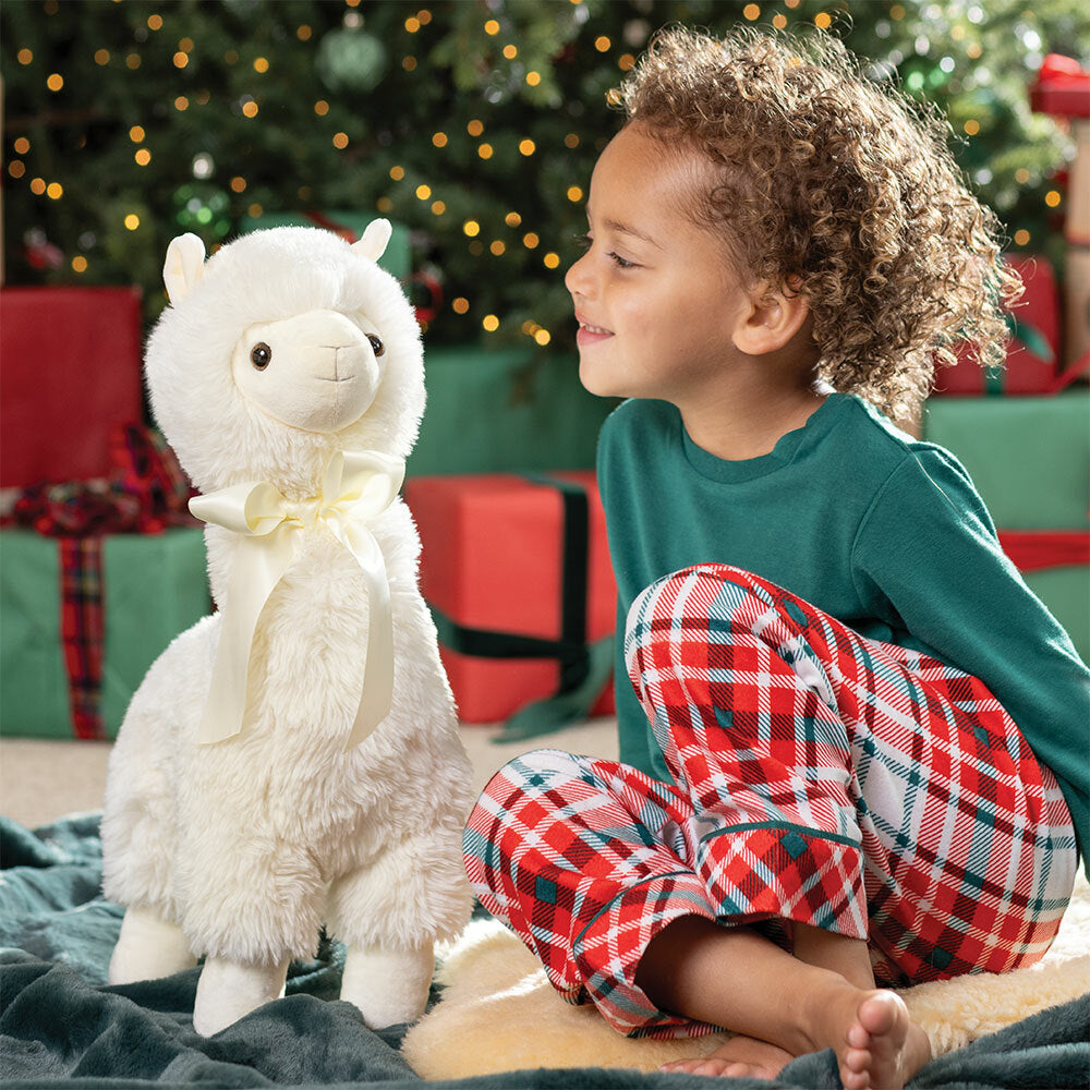 Child in pajamas sitting next to a plush llama toy in front of a decorated Christmas tree.