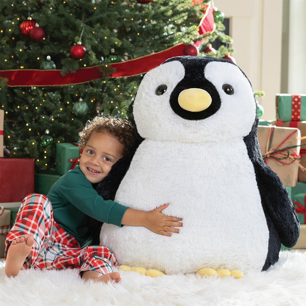 Child hugging a large plush penguin in front of a decorated Christmas tree.