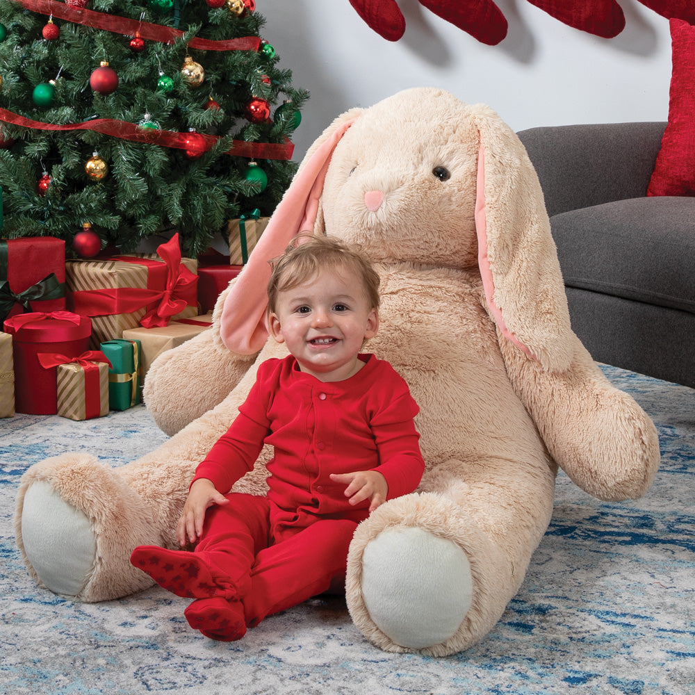 Child in red pajamas sitting on a large beige bunny plush toy with a Christmas tree and gifts in the background.