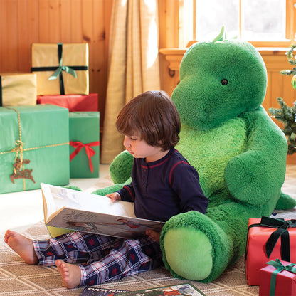 Child reading a book to a large green stuffed dinosaur surrounded by Christmas gifts.