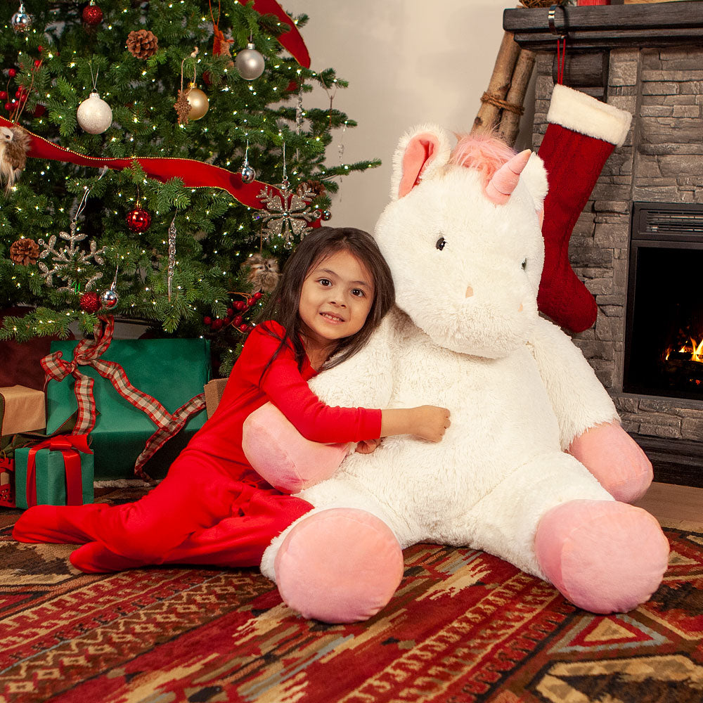 Child in red pajamas hugging a large ivory unicorn plush toy in front of  Christmas gifts.