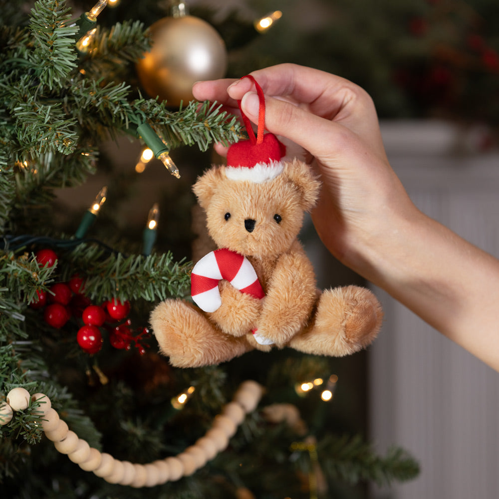 Plush teddy bear ornament wearing Santa hat and holding a red and white peppermint candy hanging on Christmas tree branch.