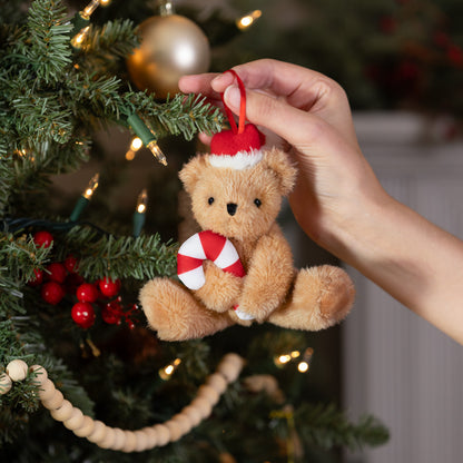Plush teddy bear ornament wearing Santa hat and holding a red and white peppermint candy hanging on Christmas tree branch.