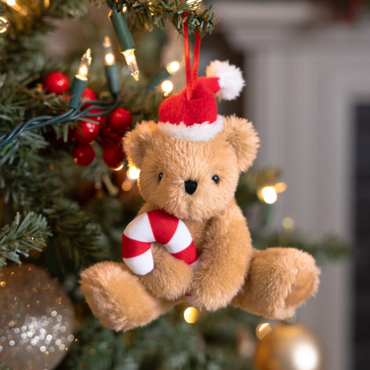 Plush teddy bear ornament wearing a Santa hat and holding a red-and-white candy cane hanging on a Christmas tree.
