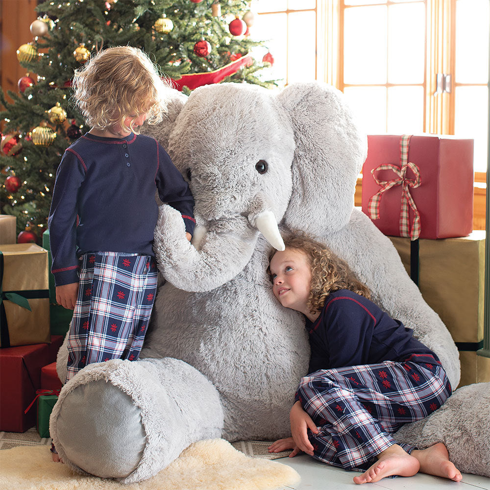 Two children playing with a large stuffed elephant in front of a decorated Christmas tree.
