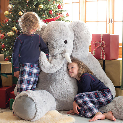 Two children playing with a large stuffed elephant in front of a decorated Christmas tree.